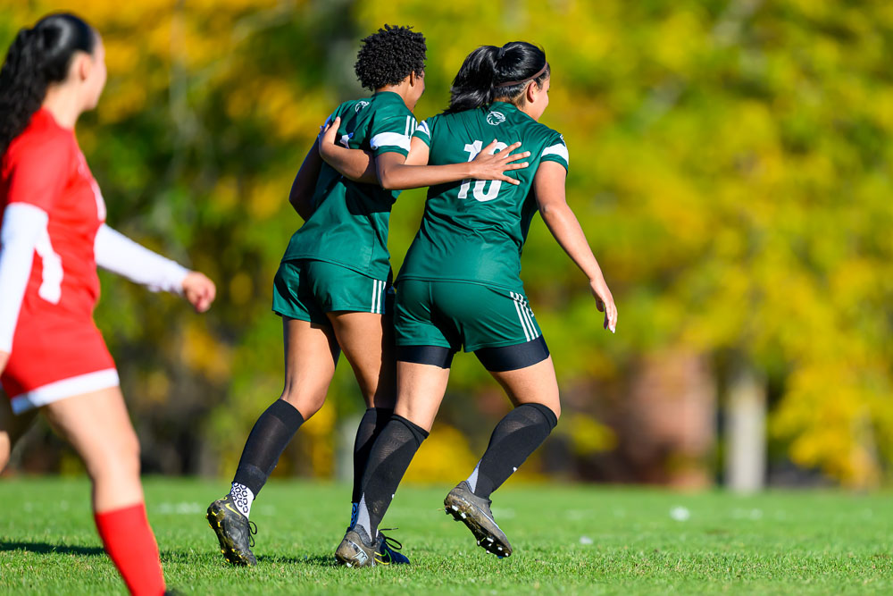 Two women with arms around each other's backs running across the soccer field
