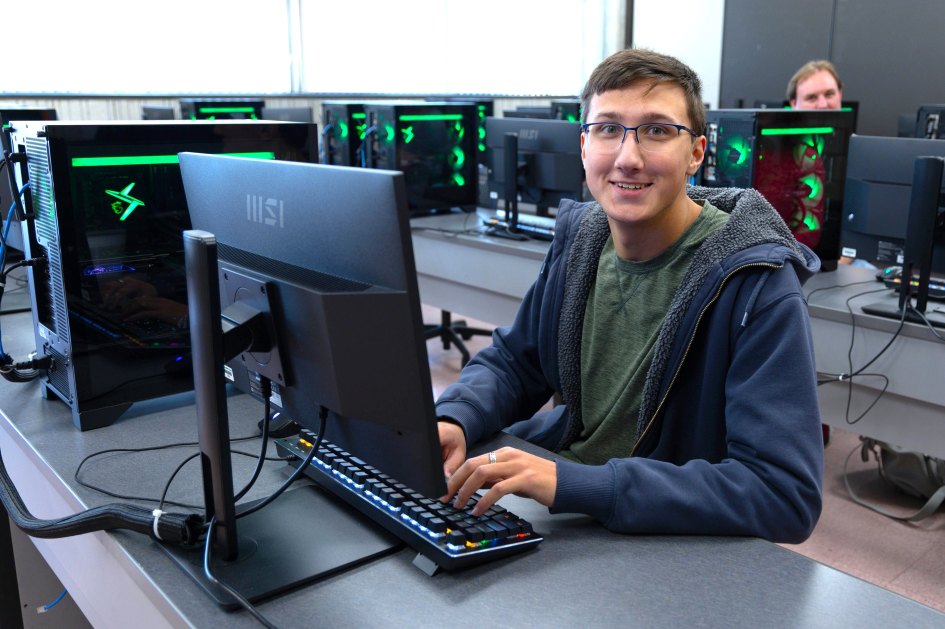 A person seated at a computer in a classroom, smiling, wearing glasses and casual clothing, surrounded by high-tech gaming PCs. The scene is bright and focused on technology.