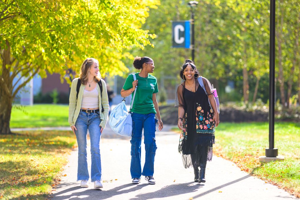 Three students walk together on a sunlit campus path in autumn, chatting and smiling. They carry bags, with vibrant green trees in the background.