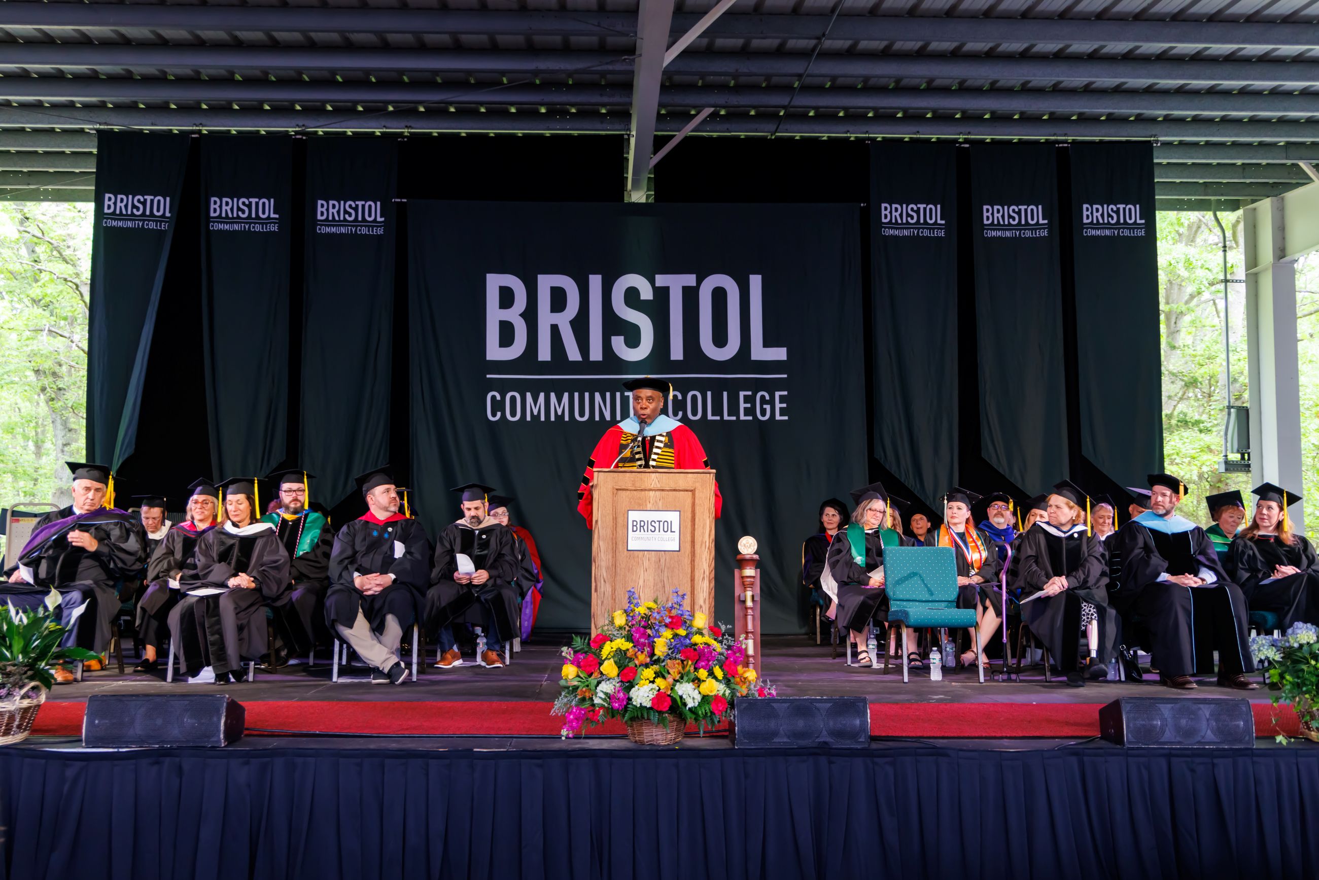 Graduation ceremony at Bristol Community College. A speaker in academic regalia stands at a podium, surrounded by faculty in caps and gowns on stage.
