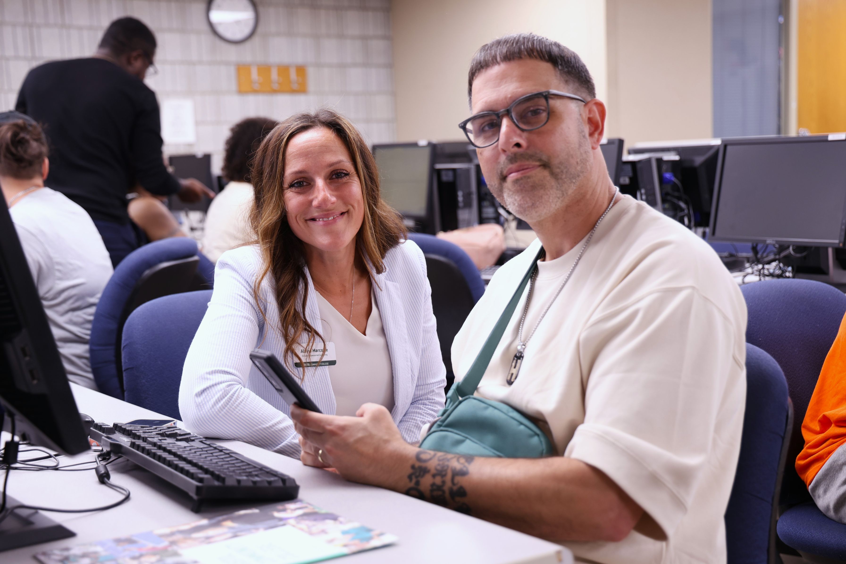 A woman in a white blazer and a man with glasses, holding a phone, sit smiling in a computer lab. The setting conveys a collaborative atmosphere.