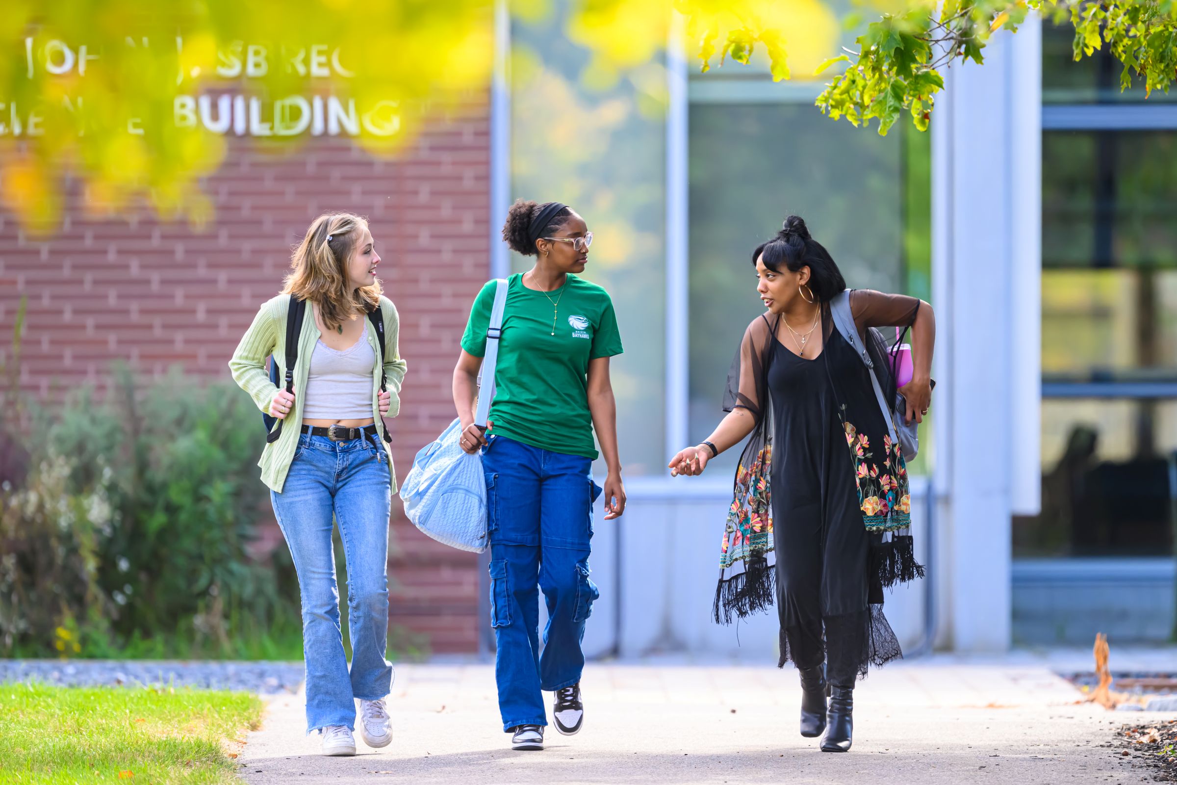 Three diverse college students walk and chat outside a brick campus building, carrying bags; sunny scene conveys friendship and connection.