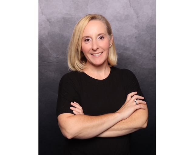 A woman with shoulder-length blonde hair smiles warmly, arms crossed, wearing a black shirt. The background is a textured gray, enhancing the portrait's professional tone.