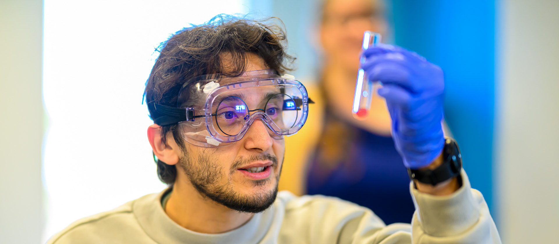Bristol Community College student wearing glasses, protective goggles, and gloves, inspecting a vial with liquid in the bottom.